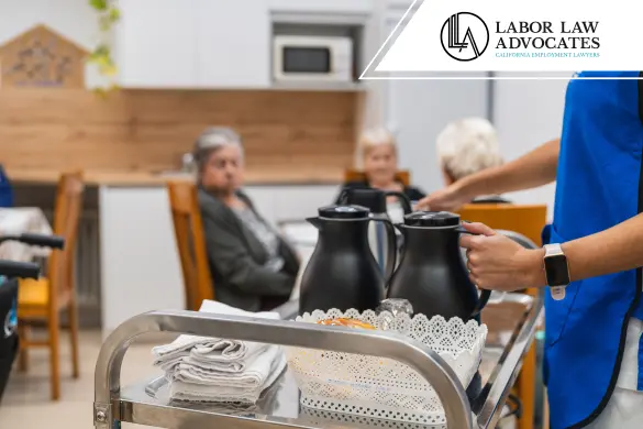 Una cuidadora con uniforme azul sirve bebidas desde un carrito a residentes mayores sentados en una mesa en un asilo de ancianos.